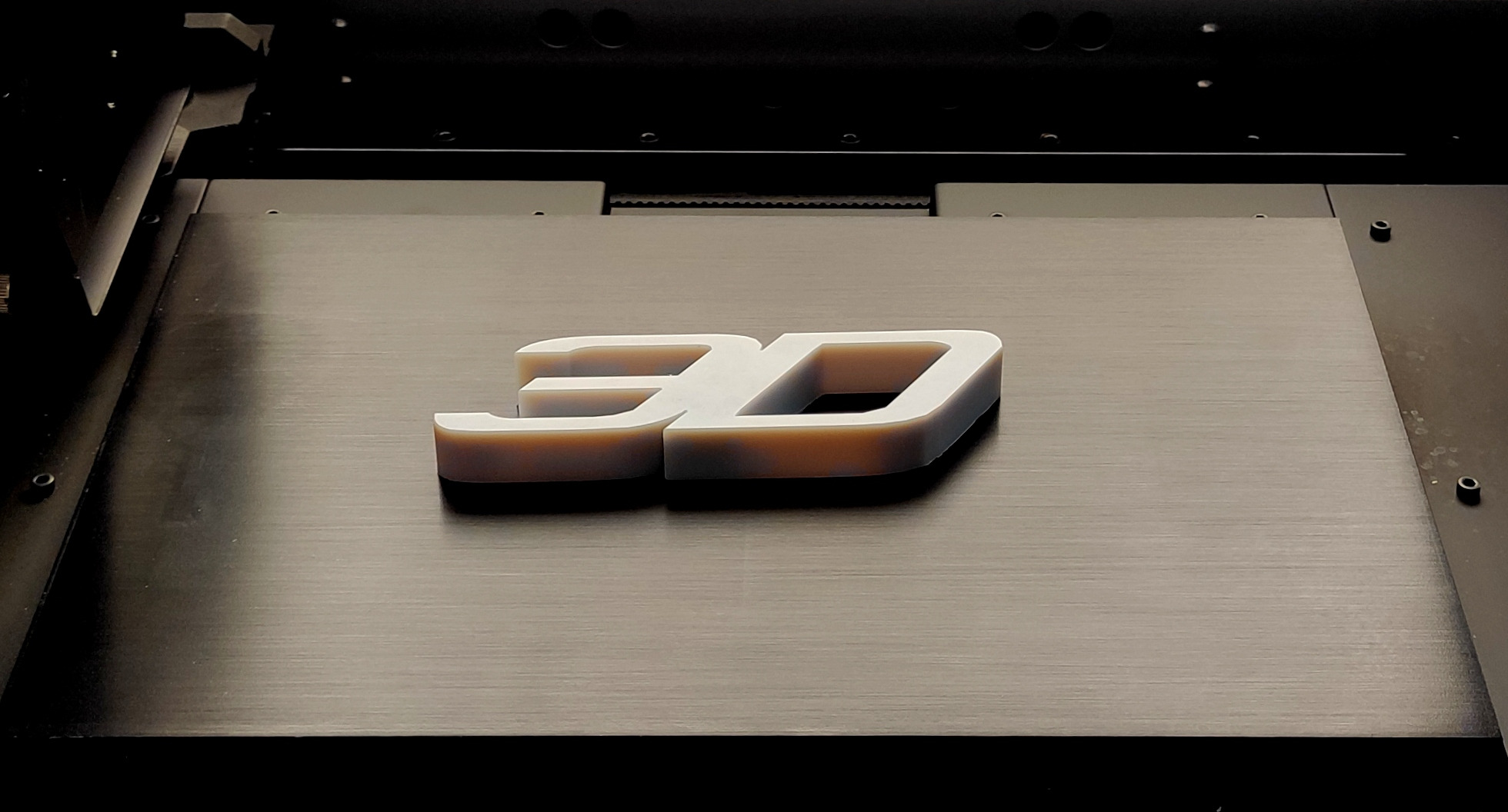 Litery 3D wydrukowane na drukarce 3D, widoczne warstwy materiału, ciemne tło drukarki.