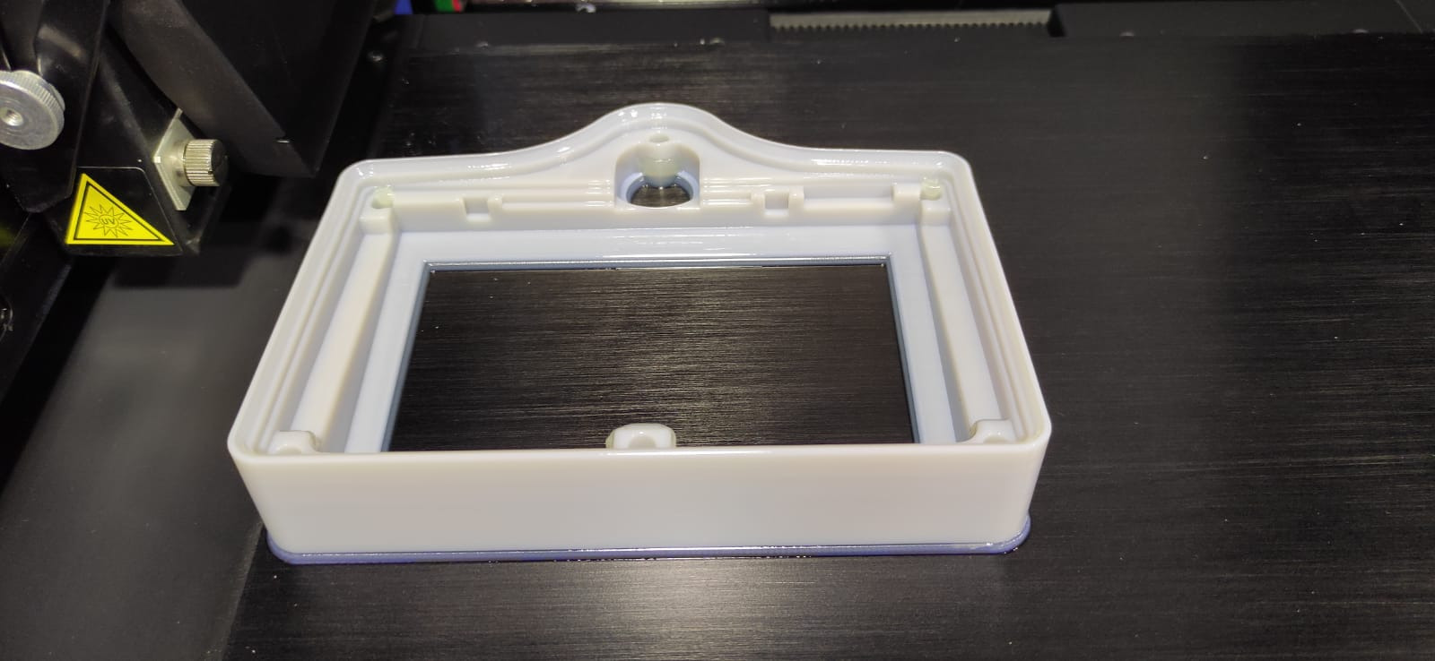 Biała, plastikowa ramka z metalową płytką wewnątrz, wydrukowana na drukarce 3D, widoczny fragment drukarki z żółtą naklejką ostrzegawczą.
