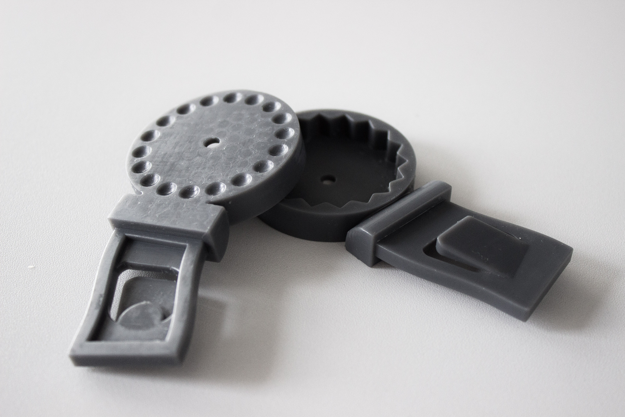 Szare, plastikowe elementy zamienne do wózka dziecięcego, wydrukowane w technologii 3D, leżące na białym tle. Widoczna okrągła część z otworami i element z wgłębieniem.