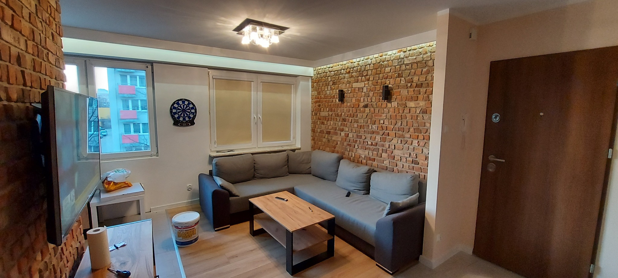 Salon po metamorfozie z ceglaną ścianą, szarym narożnikiem i drewnianym stolikiem kawowym, widok na okno z tarczą do darta i blokiem mieszkalnym za nim.