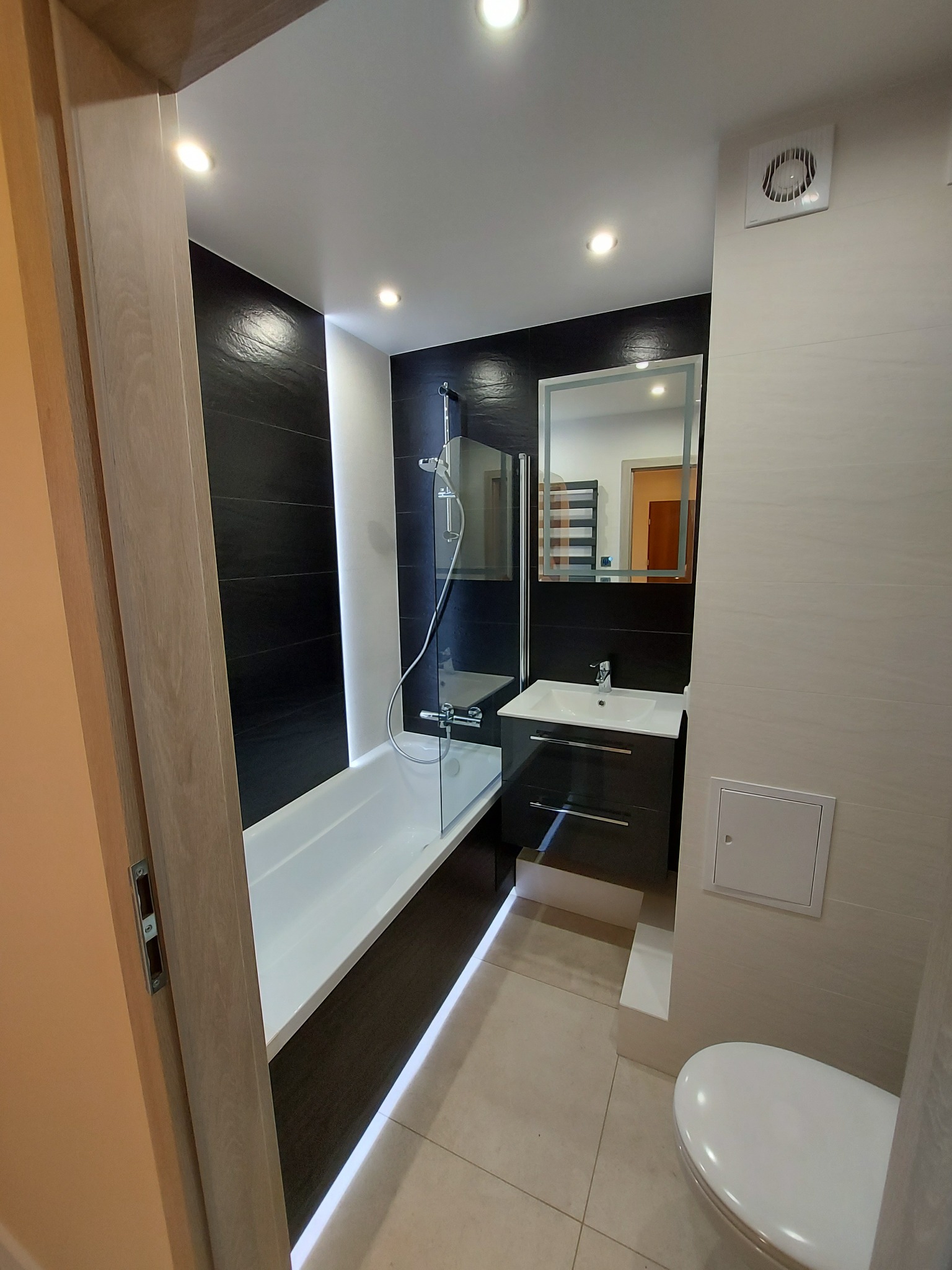 Nowoczesna łazienka z czarno-białymi płytkami, podświetlaną wanną, umywalką z szafką i lustrem z podświetleniem LED.