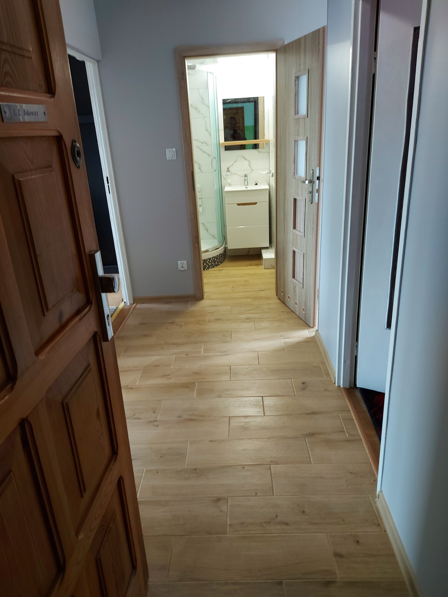 Korytarz z drewnopodobną podłogą prowadzący do łazienki z kabiną prysznicową i białą szafką pod umywalką, widok przez otwarte drzwi z drewnianą tabliczką.