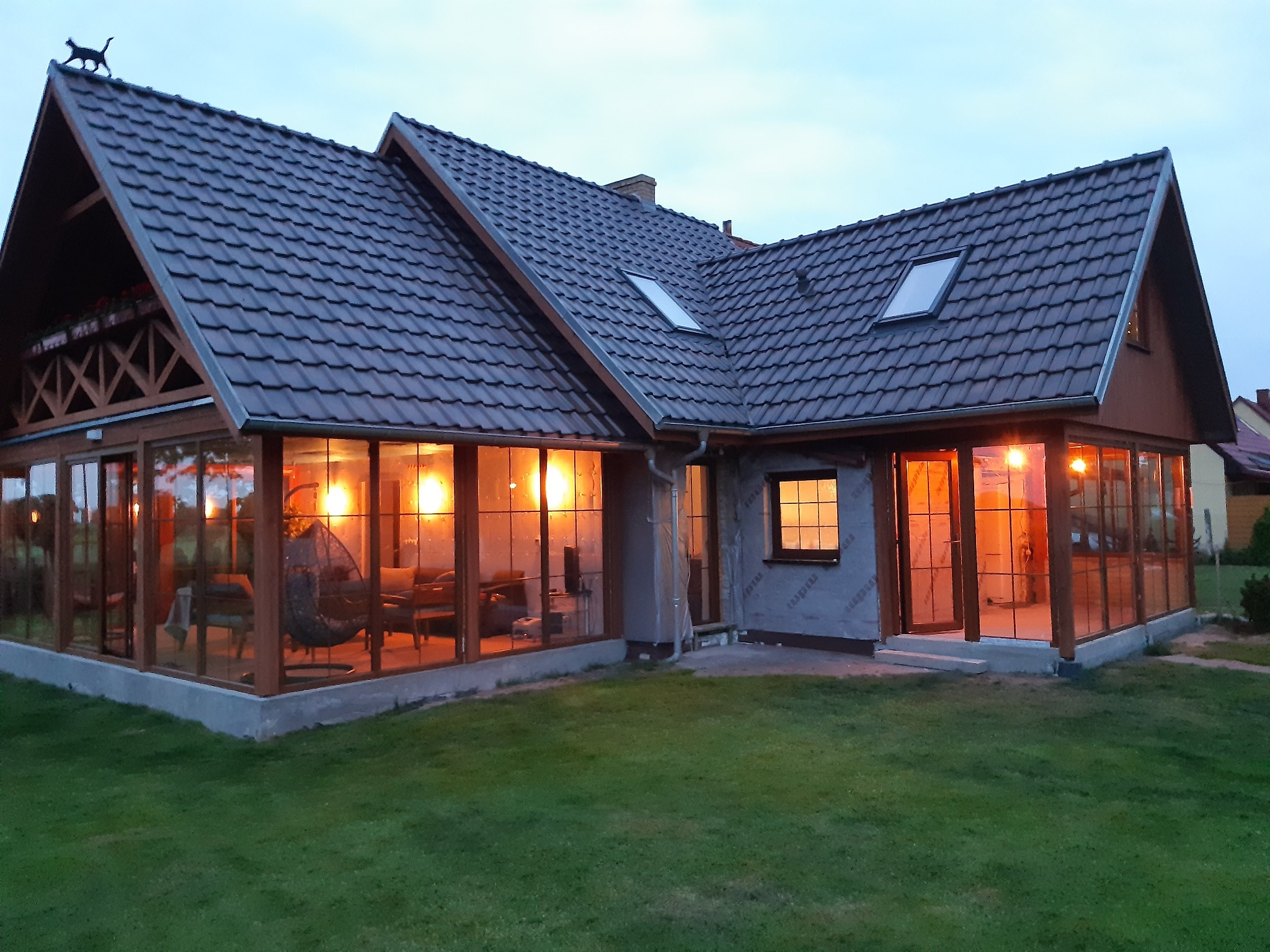 Szklana weranda z drewnianą konstrukcją dobudowana do domu, oświetlona ciepłym światłem, z widocznym wiszącym fotelem i meblami wewnątrz, na dachu figurka czarnego kota.