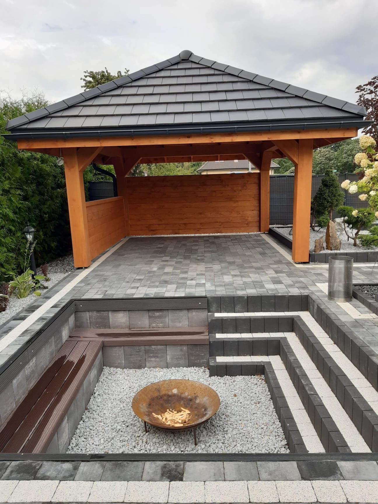 Zadaszona konstrukcja z drewnianymi słupami i ścianką z desek, wyłożona szarą kostką brukową, z zagłębieniem na palenisko wypełnionym białym żwirem i drewnianą ławką.