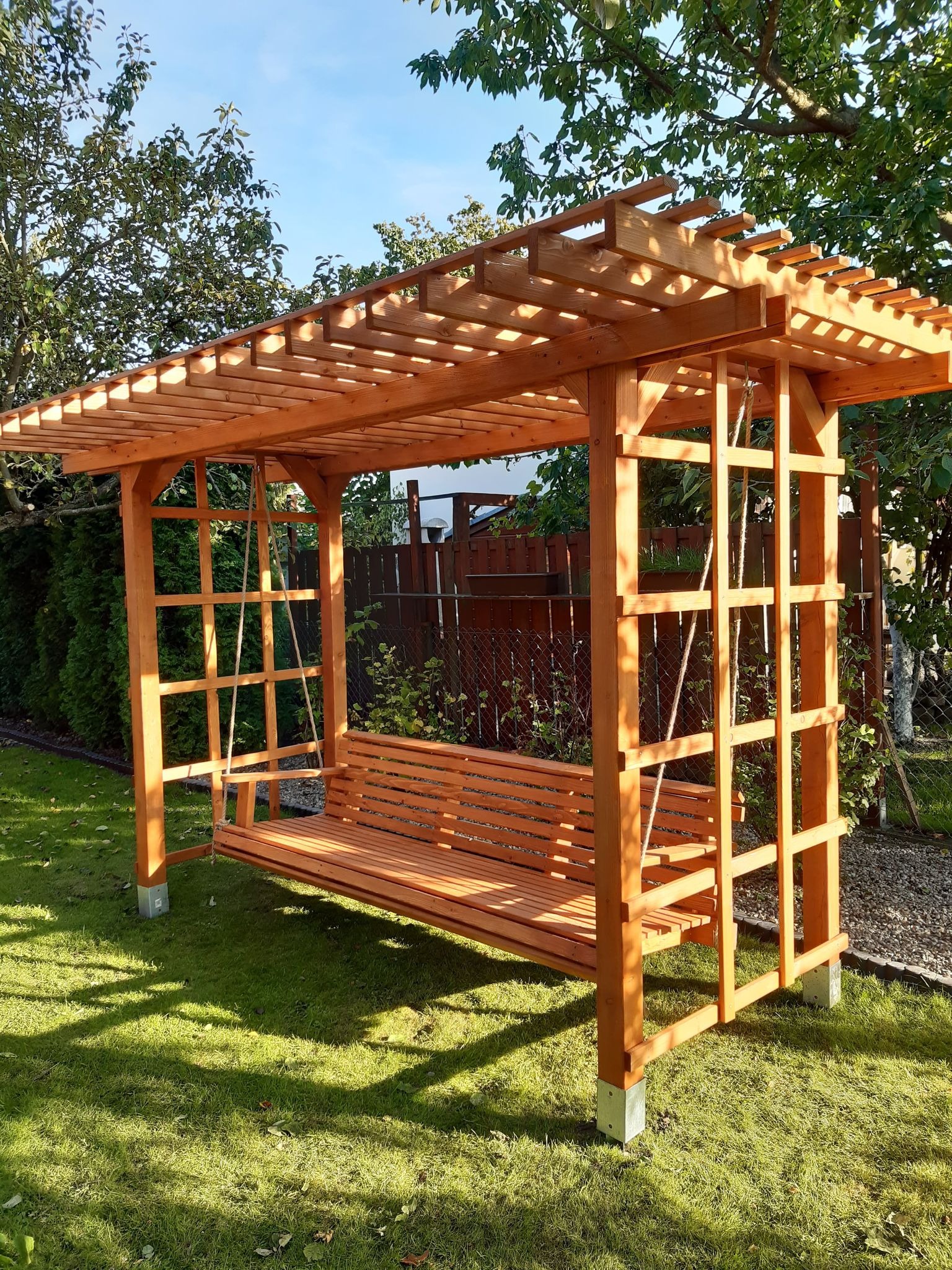 Drewniana pergola z huśtawką w ogrodzie, widok z przodu, słoneczny dzień, huśtawka zawieszona na linach.