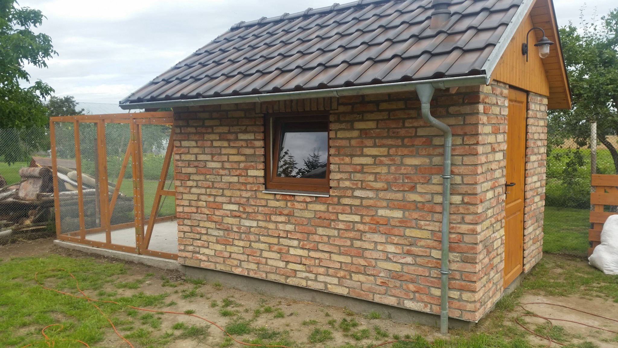 Murowana budowla z drewnianą konstrukcją dobudowaną z boku, brązowy dach z dachówki, okno z odbiciem drzew, rynna, lampa zewnętrzna, trawa wokół.