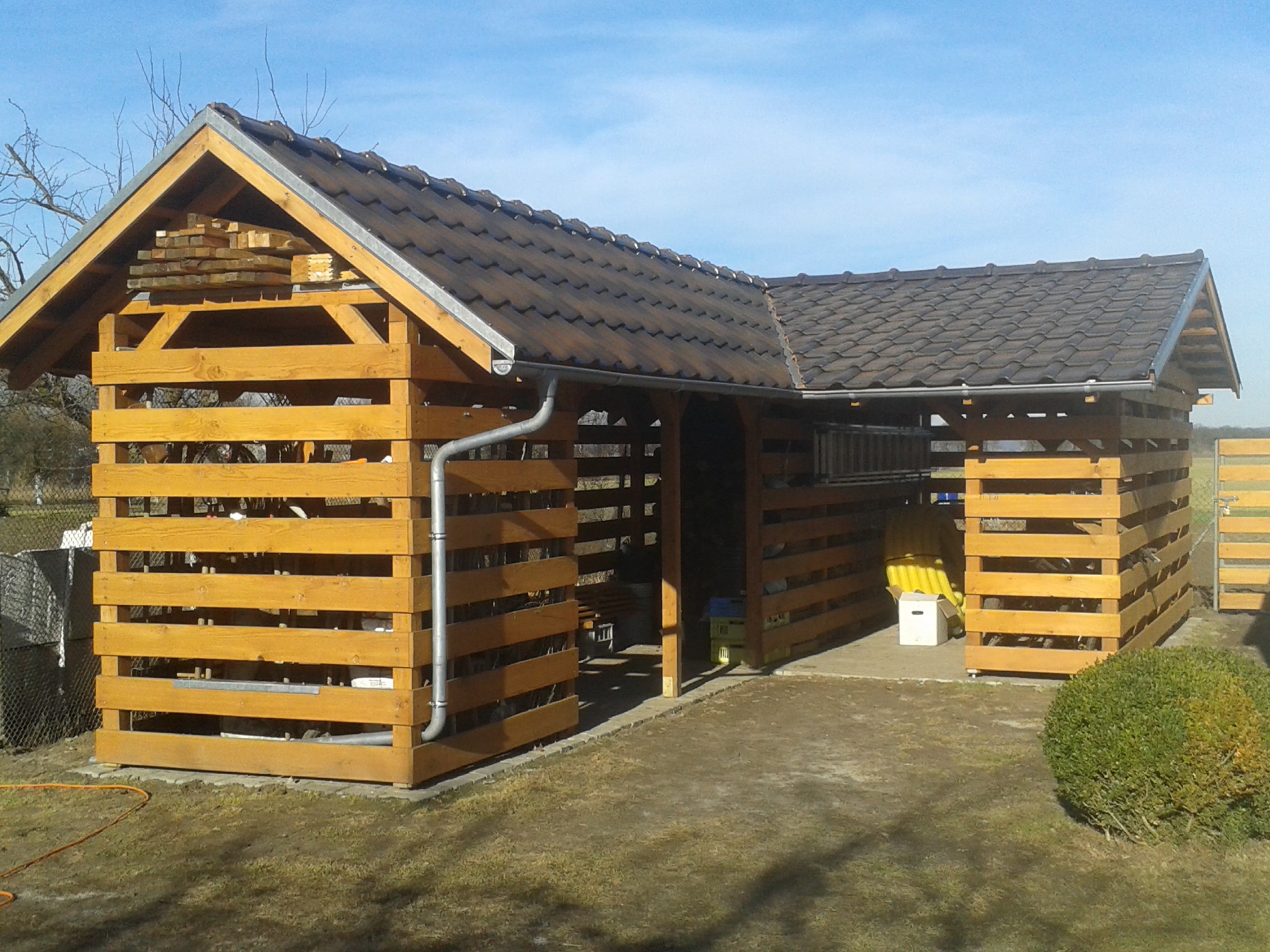 Drewniana konstrukcja z zadaszeniem z dachówki ceramicznej, potencjalnie wiata lub składzik na drewno, z widocznymi szczelinami między deskami, na zewnątrz, z elementami wyposażenia w środku.