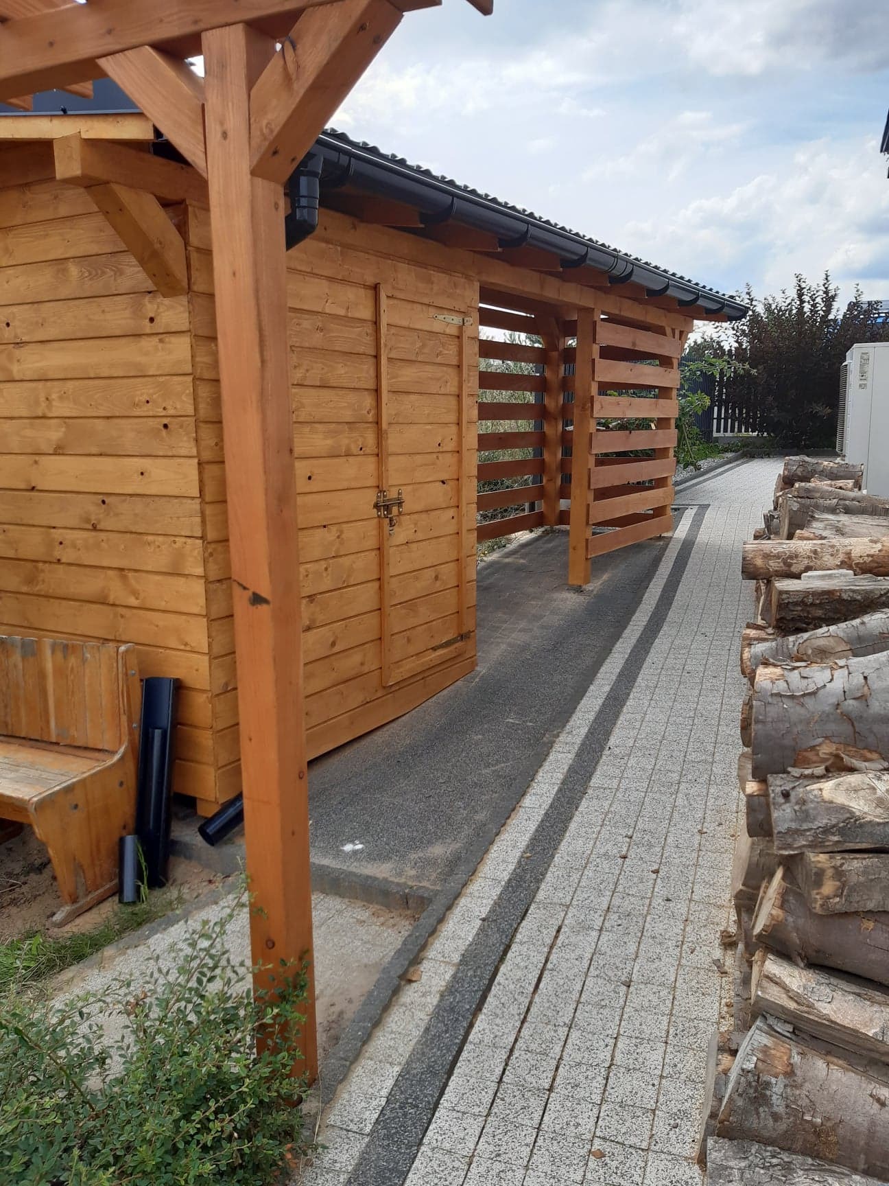 Drewniana altana z ażurowymi ścianami, widok z boku, obok ułożone drewno na opał, ścieżka z kostki brukowej.