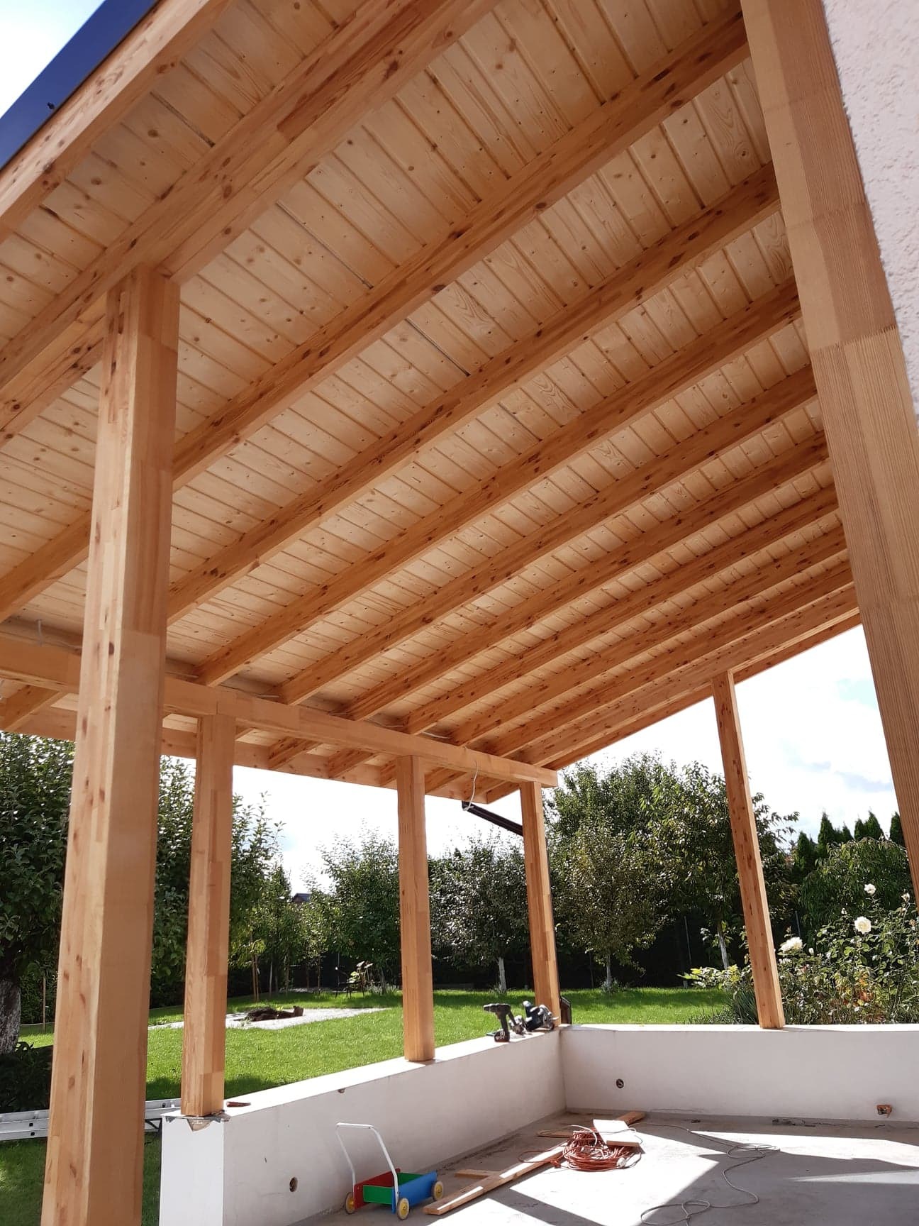 Konstrukcja z surowego drewna tworząca zadaszenie, widoczne narzędzia i zabawka dziecięca w tle ogrodowa zieleń.