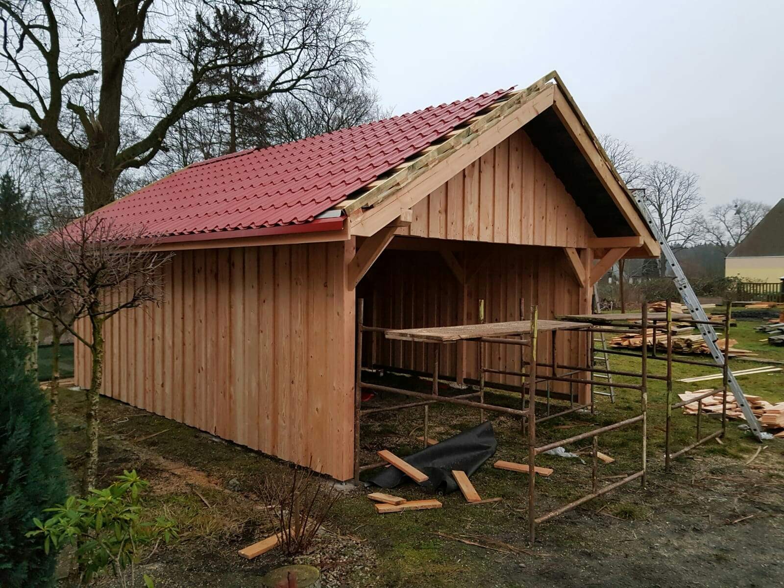 Budowa drewnianej konstrukcji z czerwonym dachem w ogrodzie, widoczne rusztowania, drabina i porozrzucane deski.