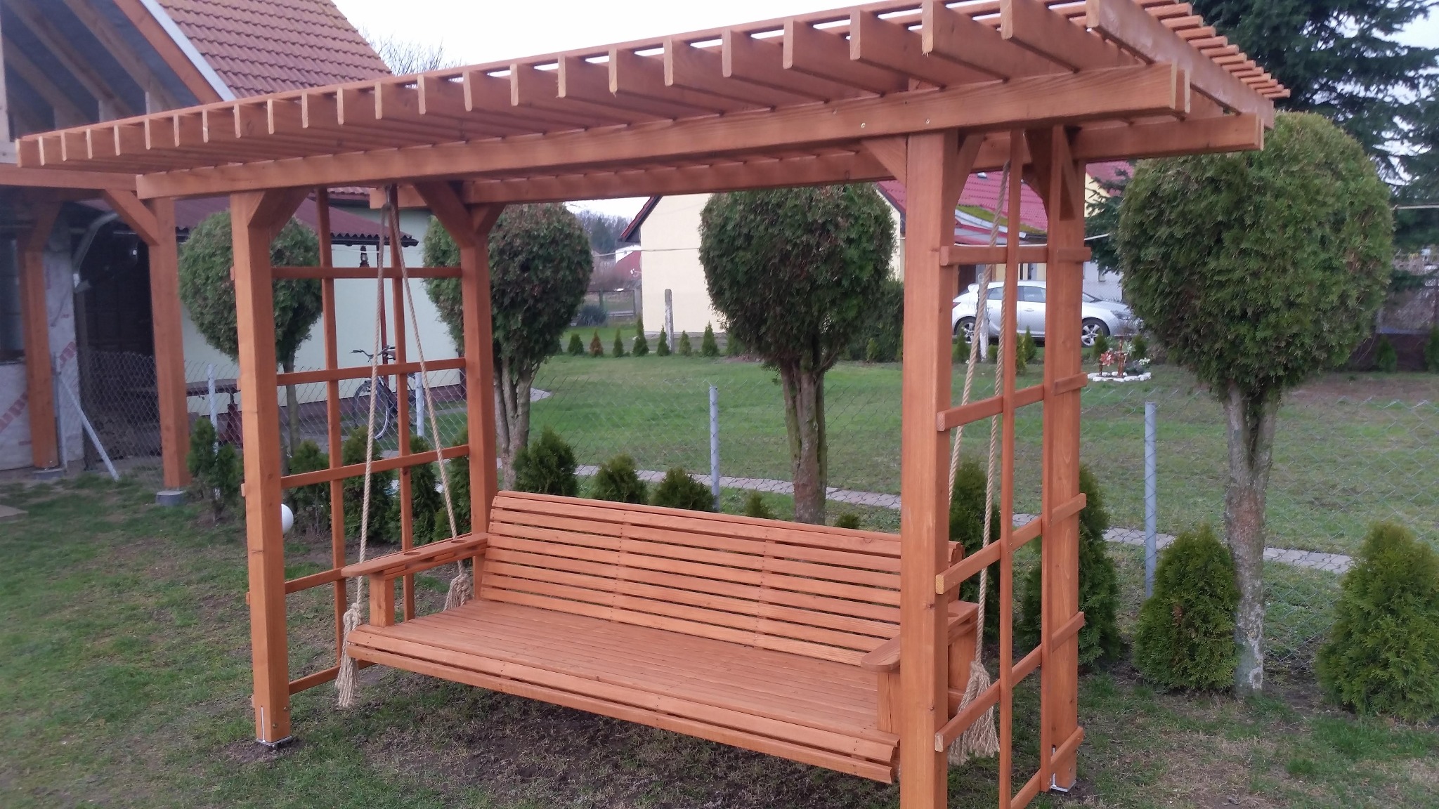 Drewniana pergola z huśtawką w ogrodzie, widok z przodu, za nią rząd tujek i siatka ogrodzeniowa.
