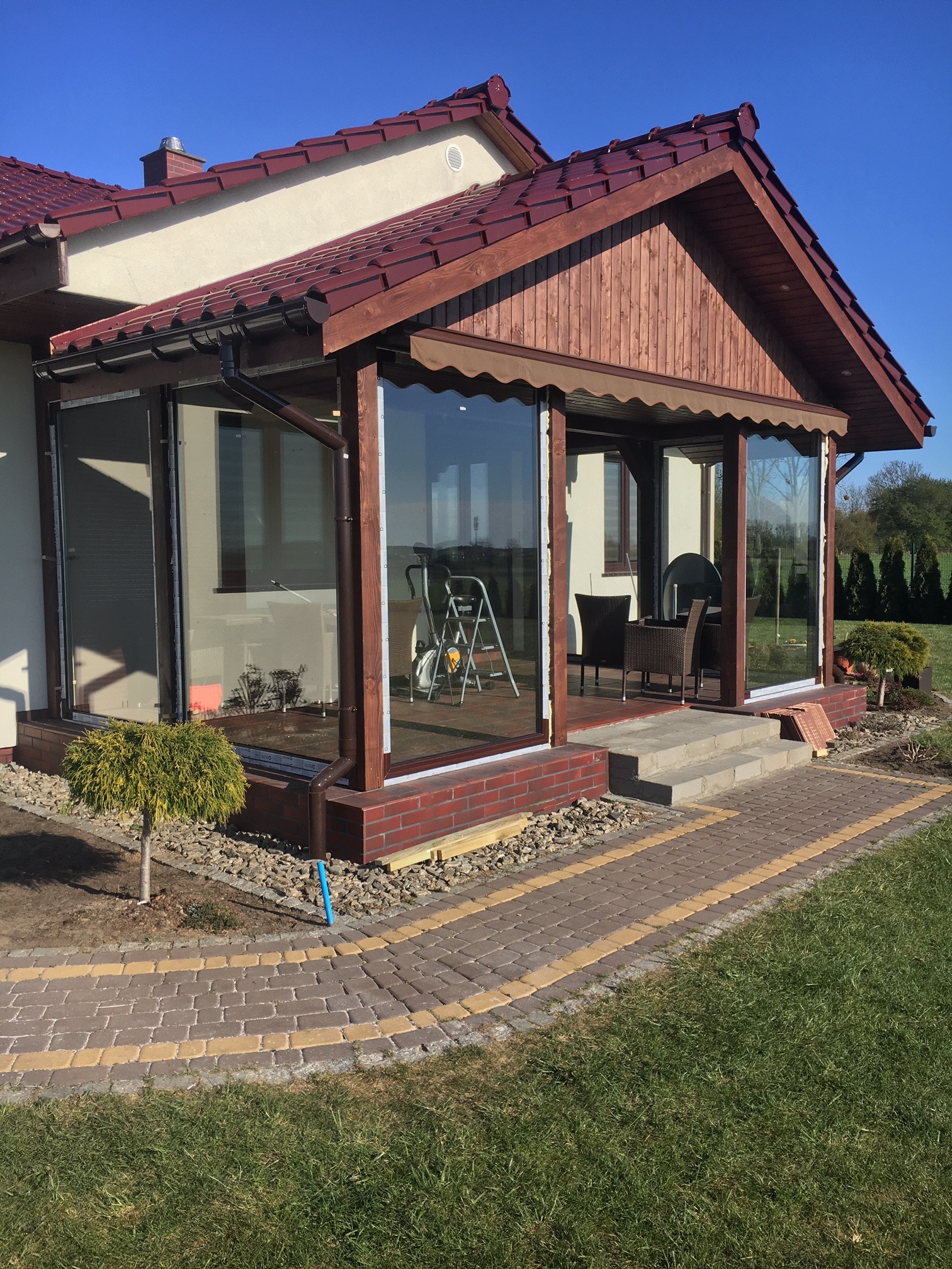 Zadaszona weranda z dużymi oknami, drewnianą konstrukcją i ceglaną podstawą, z widocznymi schodami wejściowymi i ścieżką z kostki brukowej.