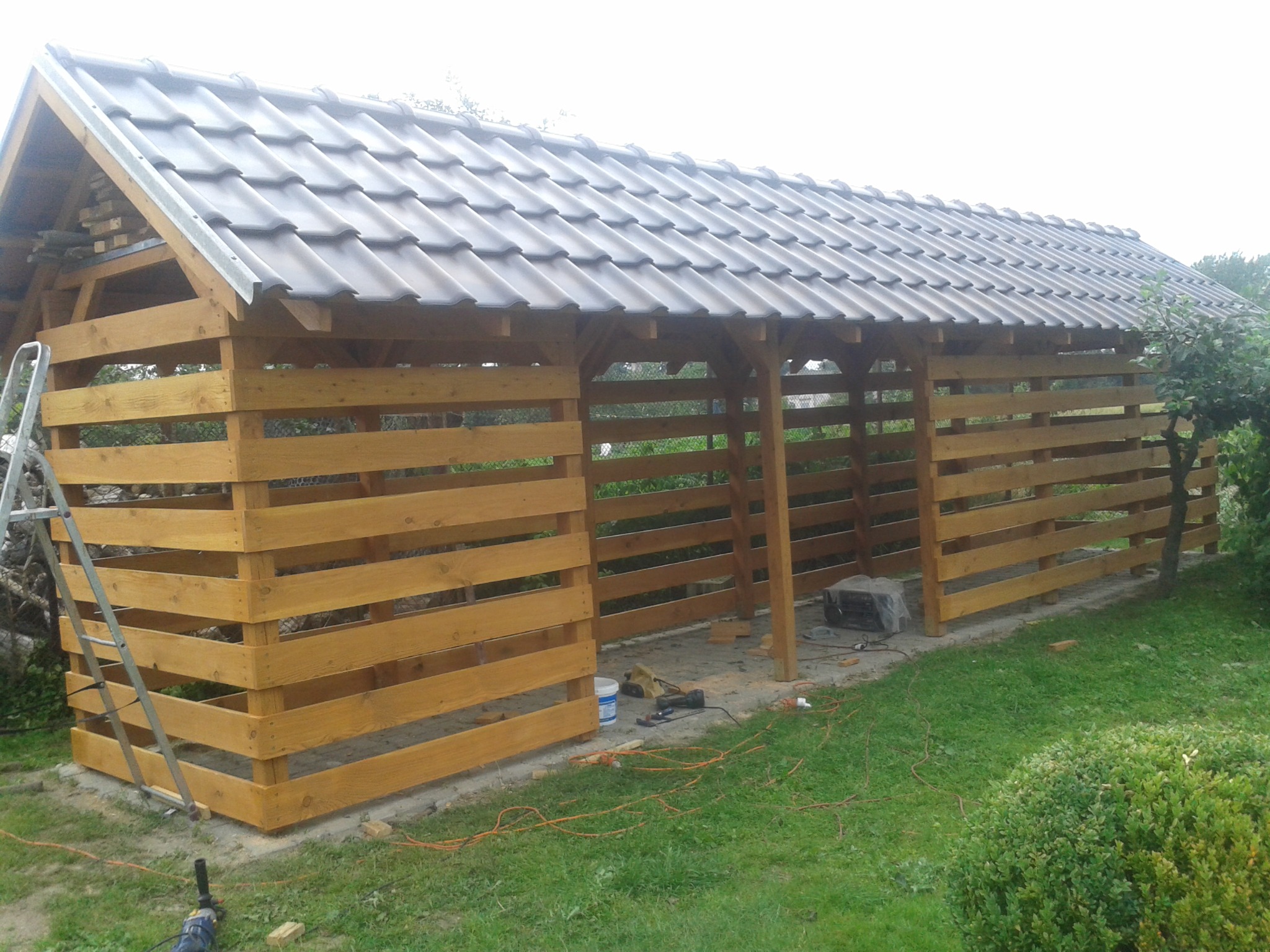 Drewniana konstrukcja z szarą dachówką w trakcie budowy, widoczne narzędzia i drabina oparta o ścianę, otoczenie zielone.