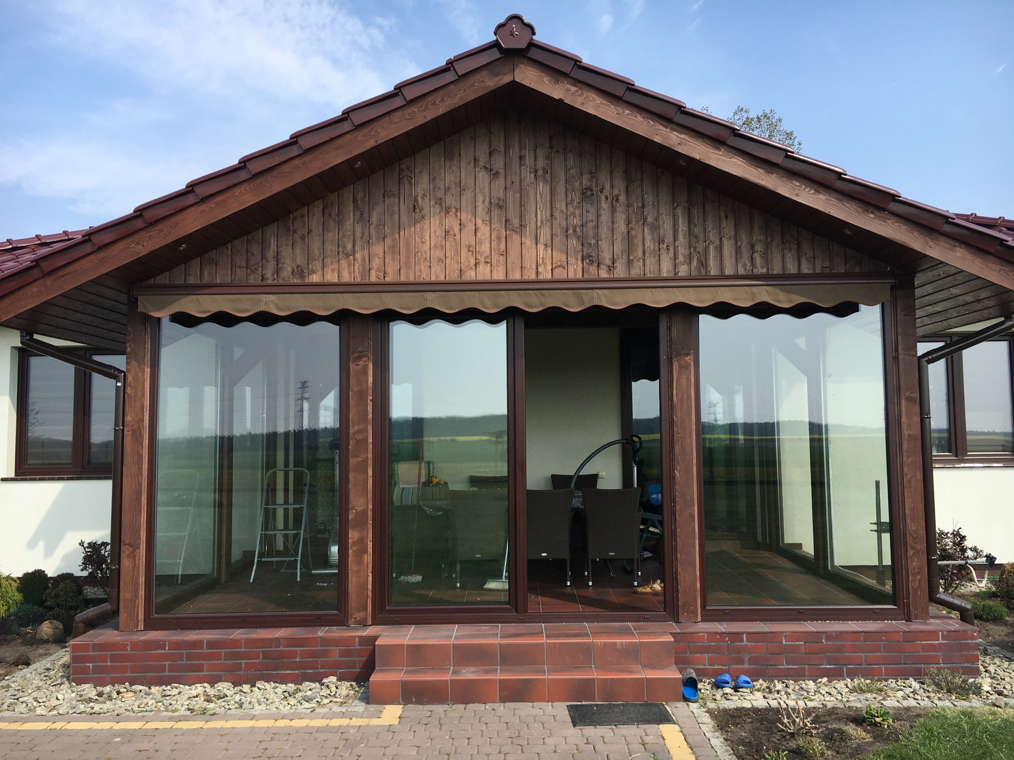 Zadaszona konstrukcja z drewna i szkła z brązowymi ramami, z widocznymi schodami z cegły i fragmentem ogrodu z kostki brukowej, wewnątrz widoczne krzesła i drabina.