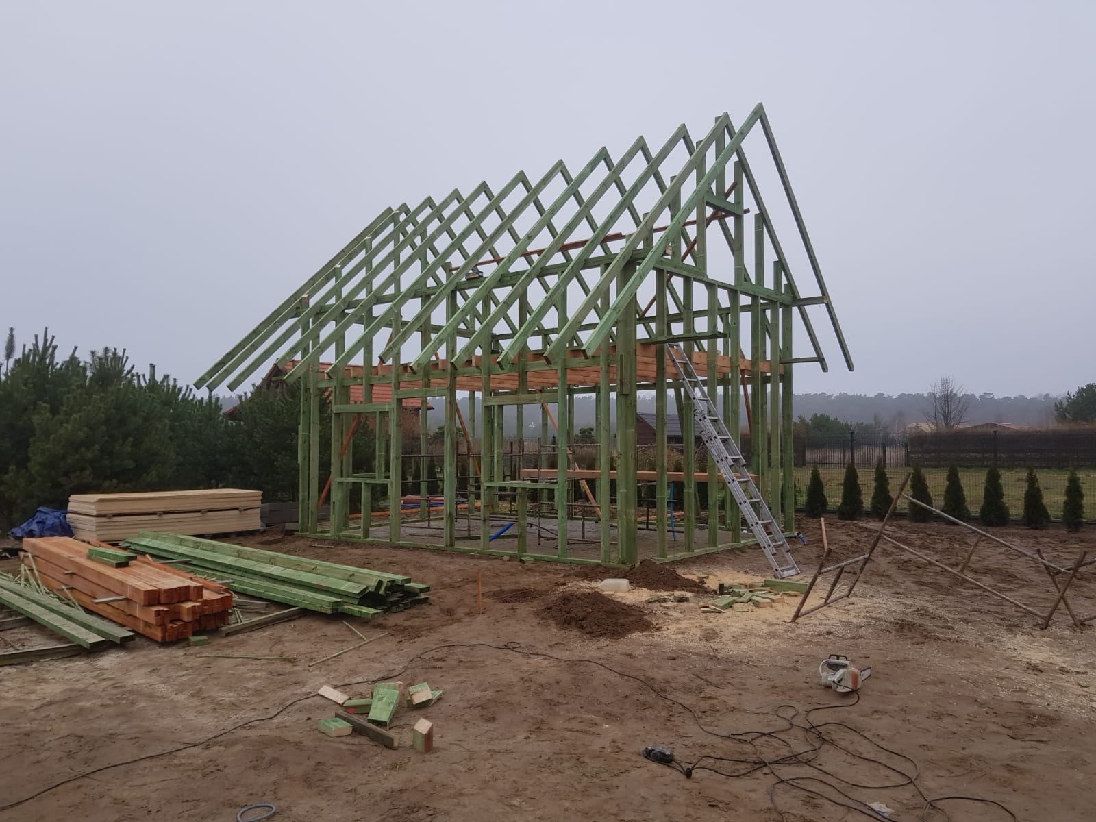 Konstrukcja drewniana w trakcie budowy, widoczne elementy szkieletu i więźby dachowej, drabina oparta o ścianę, deski i narzędzia na placu budowy, pochmurny dzień.