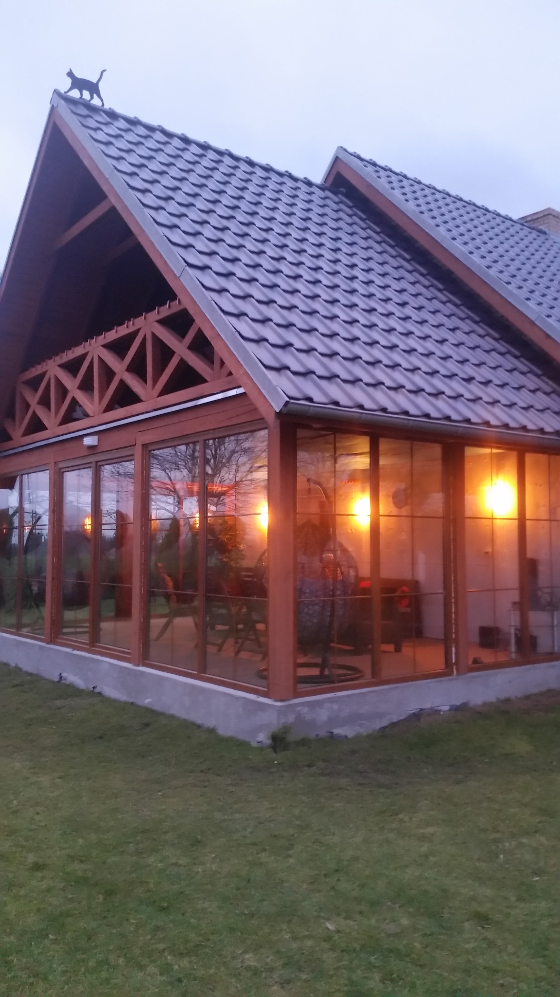 Przeszklona konstrukcja z drewnianą ramą, prawdopodobnie weranda lub oranżeria, z oświetlonym wnętrzem i dekoracją w postaci sylwetki kota na szczycie dachu.