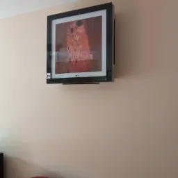 Klimatyzator LG o mocy 3,5 kW w czarnej obudowie, zamontowany na beżowej ścianie, prezentujący obraz Gustava Klimta 'Pocałunek', widoczny fragment czerwonej kanapy.