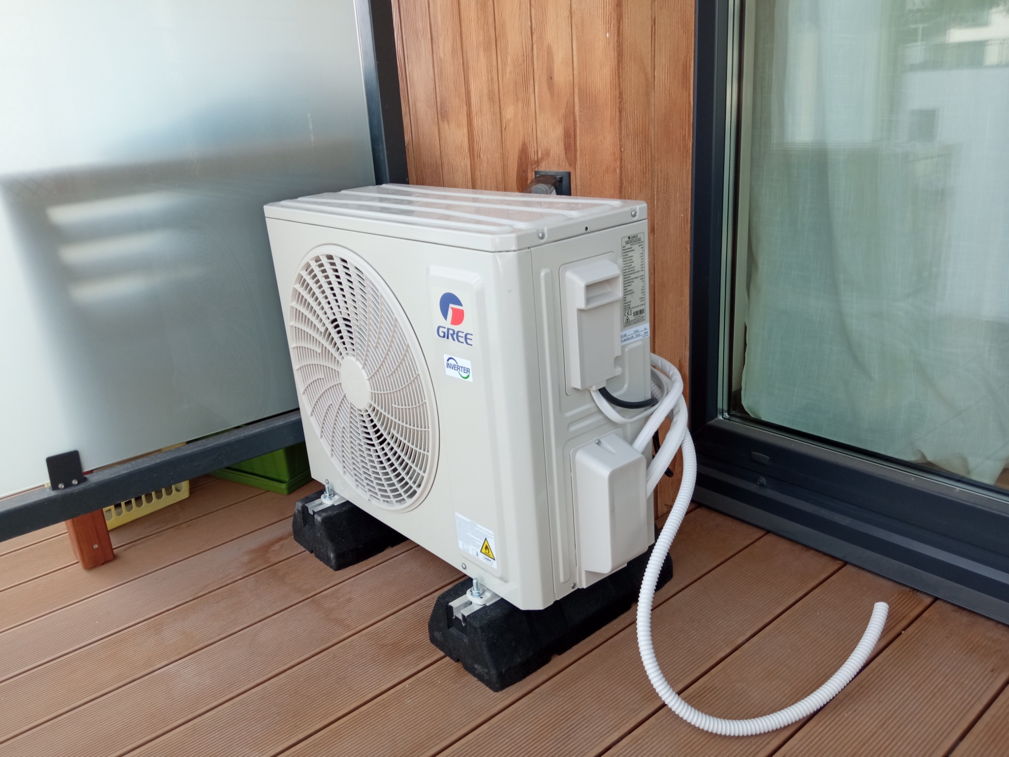 Zewnętrzna jednostka klimatyzacji marki Gree zamontowana na czarnych podkładkach na drewnianym tarasie, z widocznymi przewodami i ścianą z drewnianych paneli w tle.