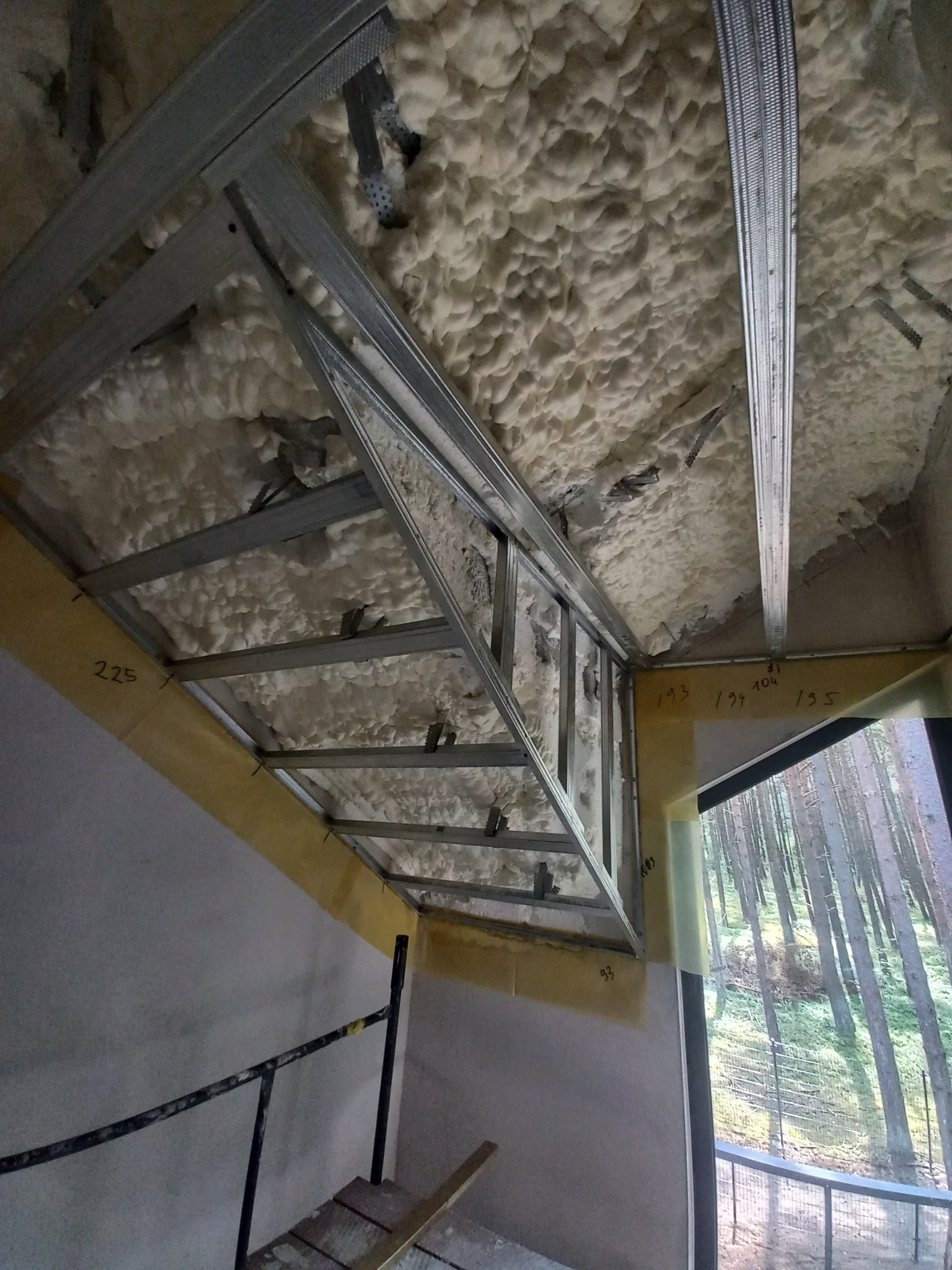 Montaż metalowej konstrukcji sufitu podwieszanego z izolacją piankową w Kołobrzegu, widok z dołu na ruszt i pianę, z oknem z widokiem na las w tle.