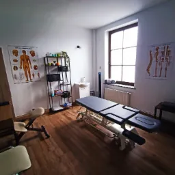 Jasne wnętrze gabinetu fizjoterapii z regulowanym stołem do masażu, regałem z dekoracjami i plakatami anatomicznymi na ścianach.