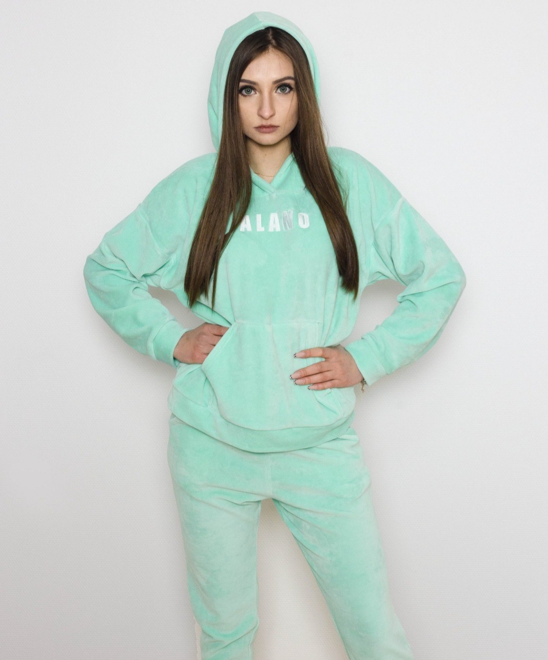 Kobieta w miętowym welurowym dresie z kapturem, ręce oparte na biodrach, na białym tle.