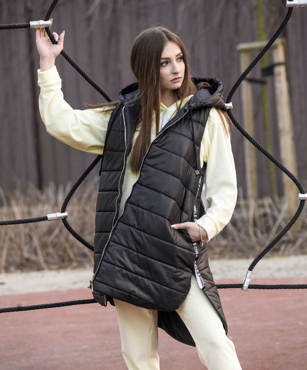 Portret młodej kobiety w czarnej, pikowanej kamizelce z kapturem, pozującej na tle konstrukcji linowej na placu zabaw. Ubrana w pastelowy dres, z ręką opartą o linę i wzrokiem skierowanym w bok.