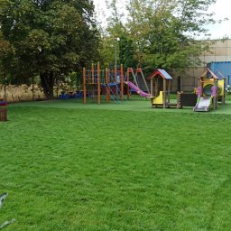 SIŁA W OGRODZIE SPÓŁKA Z OGRANICZONĄ ODPOWIEDZIALNOŚCIĄ - Zielony plac zabaw z syntetyczną trawą, kolorowymi konstrukcjami do zabawy i piknikowymi stolikami, otoczony drzewami i ogrodzeniem.