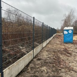SIŁA W OGRODZIE SPÓŁKA Z OGRANICZONĄ ODPOWIEDZIALNOŚCIĄ - Długi, szary metalowy płot panelowy z betonową podmurówką, ciągnący się wzdłuż terenu. W tle widoczne krzewy i drzewa bez liści oraz niebieski przenośny WC.