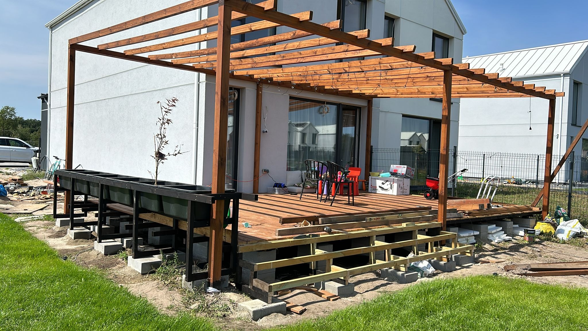 Drewniana konstrukcja pergoli w trakcie budowy, z widocznymi elementami tarasu i donicami. Ujęcie z perspektywy trawnika, w tle nowoczesny dom.