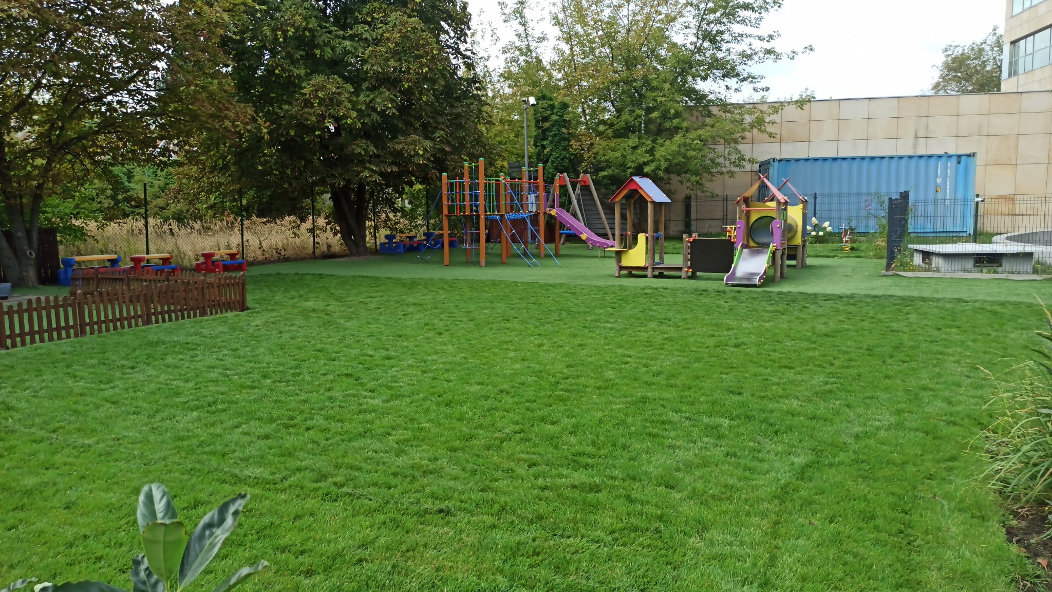 Zielony plac zabaw z syntetyczną trawą, kolorowymi konstrukcjami do zabawy i piknikowymi stolikami, otoczony drzewami i ogrodzeniem.