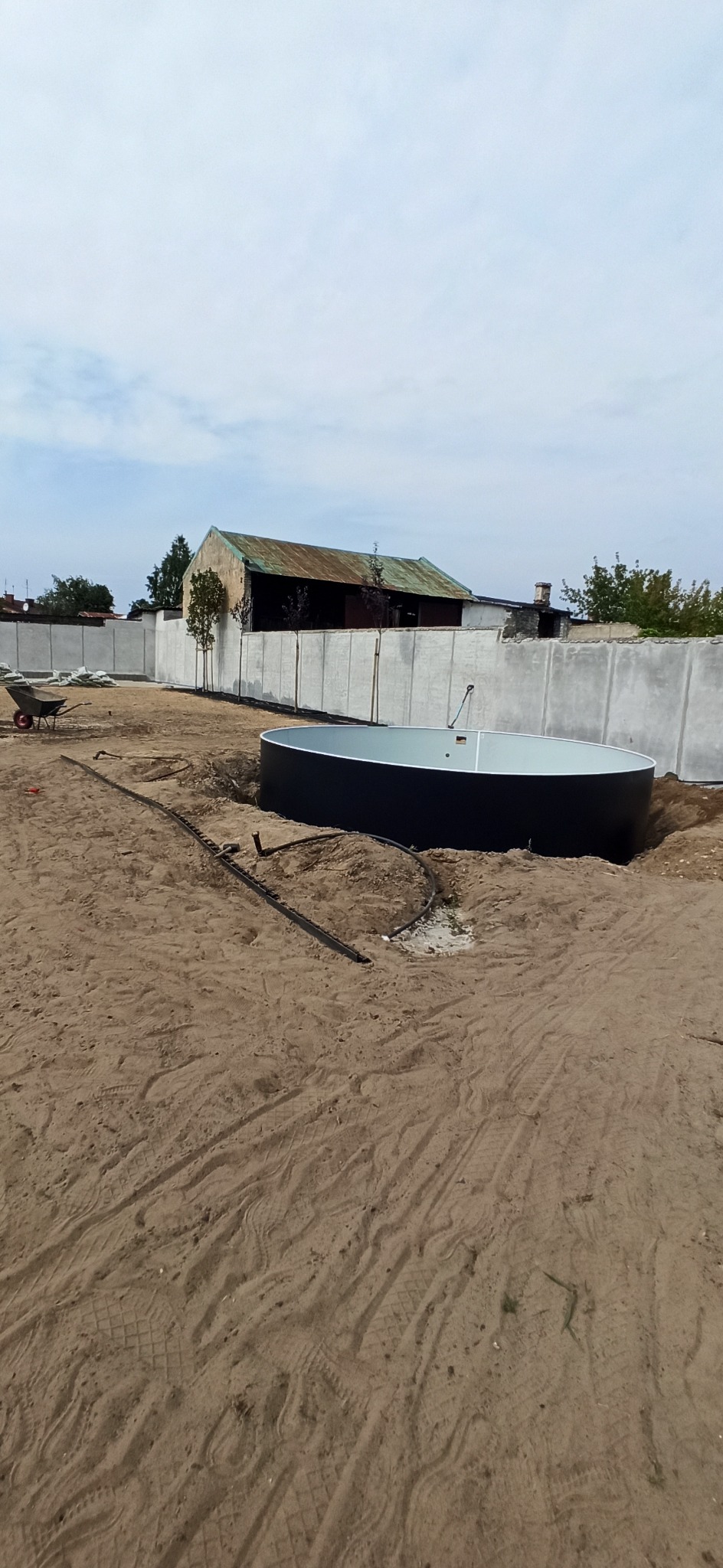Okrągły, czarny zbiornik na wodę w wykopanym dole, z widocznymi rurami i piaskiem. W tle szary betonowy płot i budynek gospodarczy.