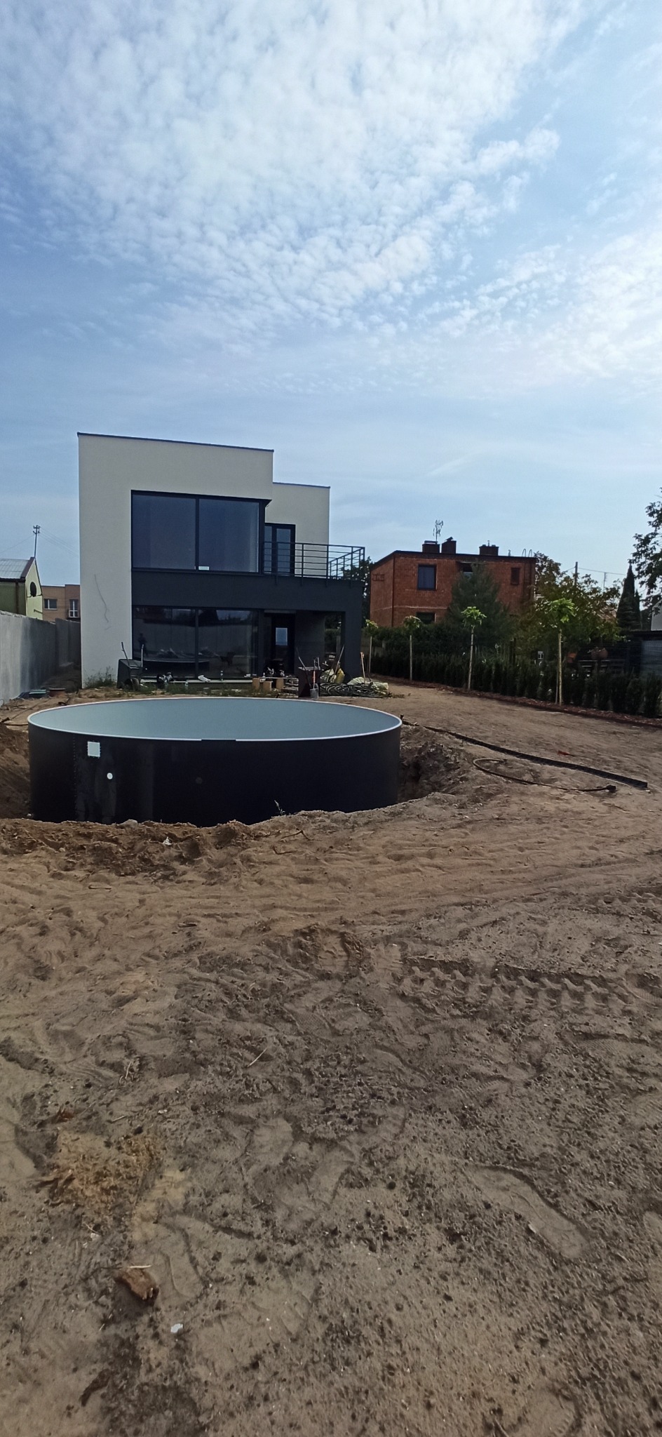 Montaż okrągłego basenu w ogrodzie nowoczesnego domu. Widoczne przygotowane podłoże i częściowo zagospodarowany teren. Błękitne niebo z chmurami w tle.
