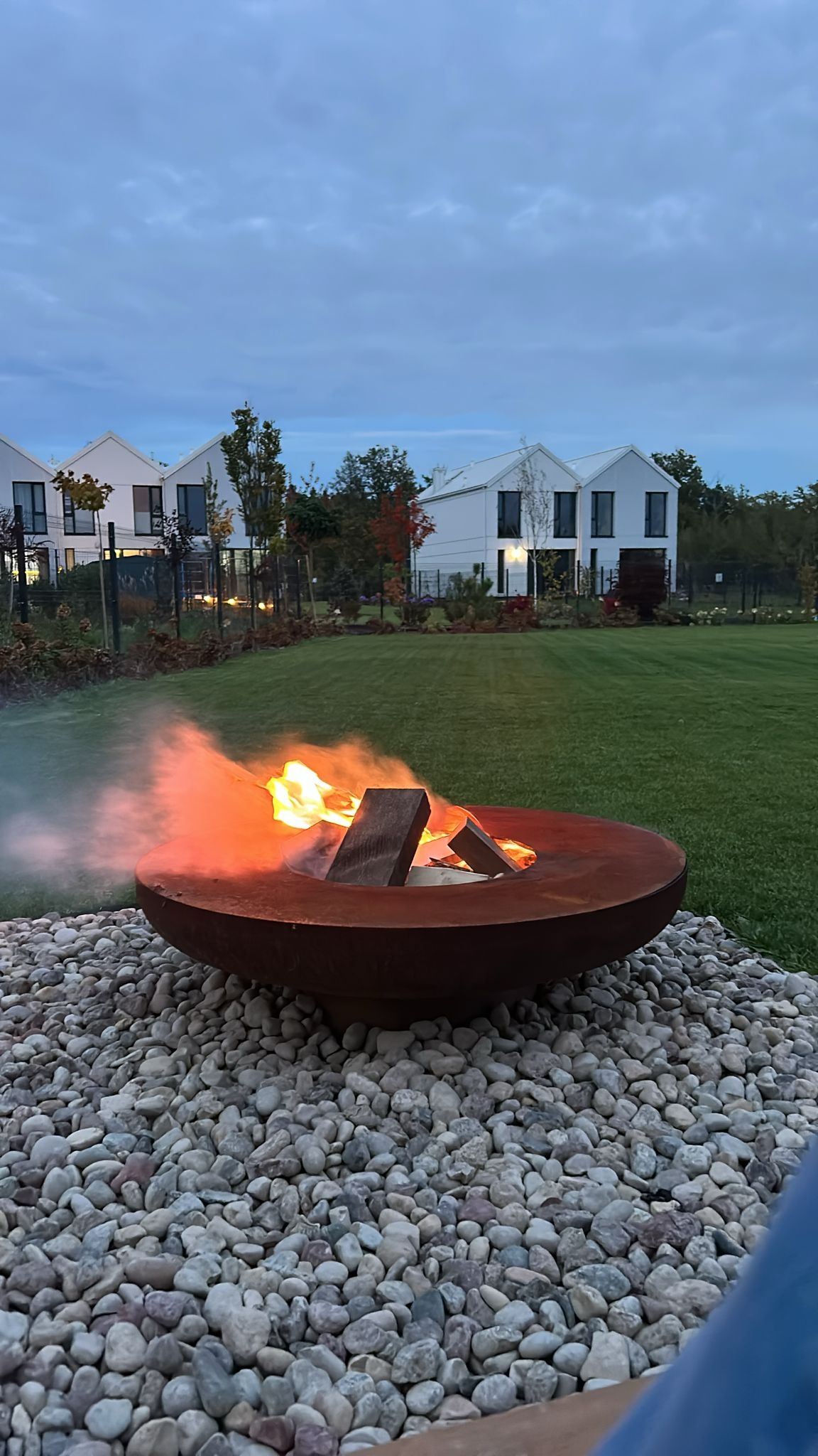 Ognisko w metalowej misie na kamiennym podłożu, w tle trawnik i nowoczesne domy. Wieczorny krajobraz ogrodu z elementami dekoracyjnymi.