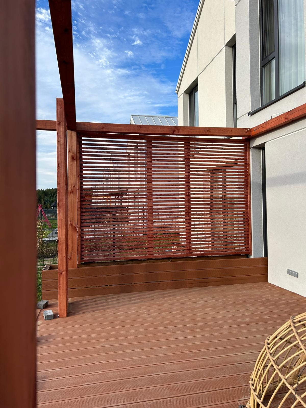 Nowoczesna drewniana pergola z poziomymi listwami, tworząca zacienioną przestrzeń na tarasie przy budynku. Widoczne elementy konstrukcyjne i deski tarasowe.