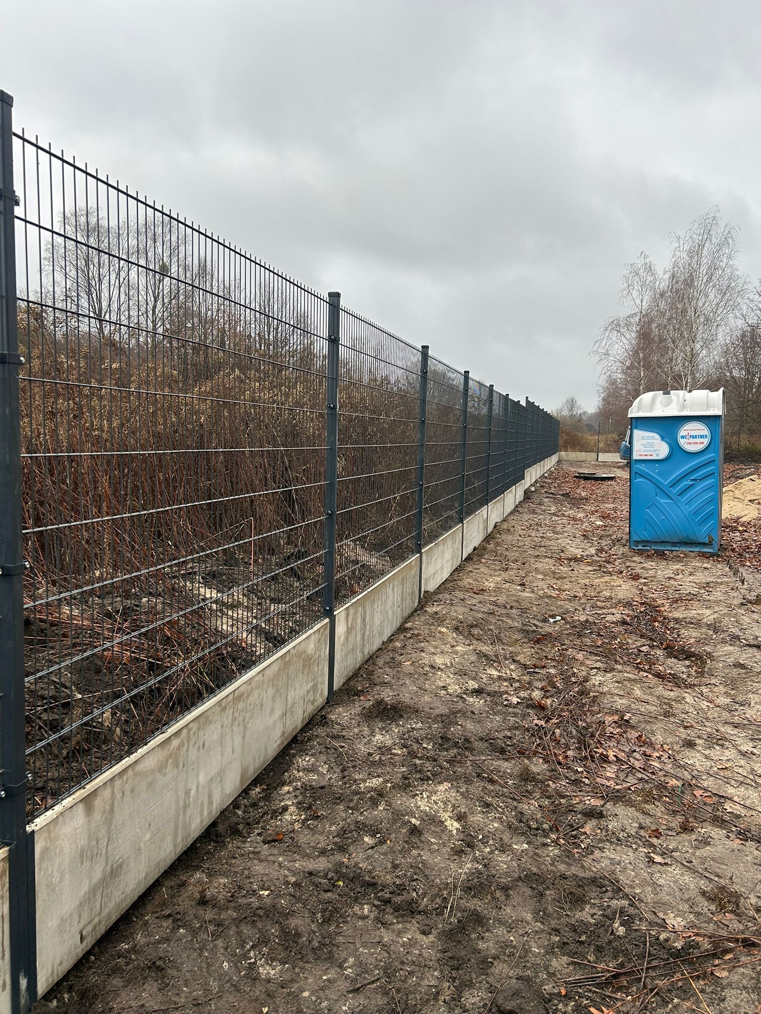Długi, szary metalowy płot panelowy z betonową podmurówką, ciągnący się wzdłuż terenu. W tle widoczne krzewy i drzewa bez liści oraz niebieski przenośny WC.