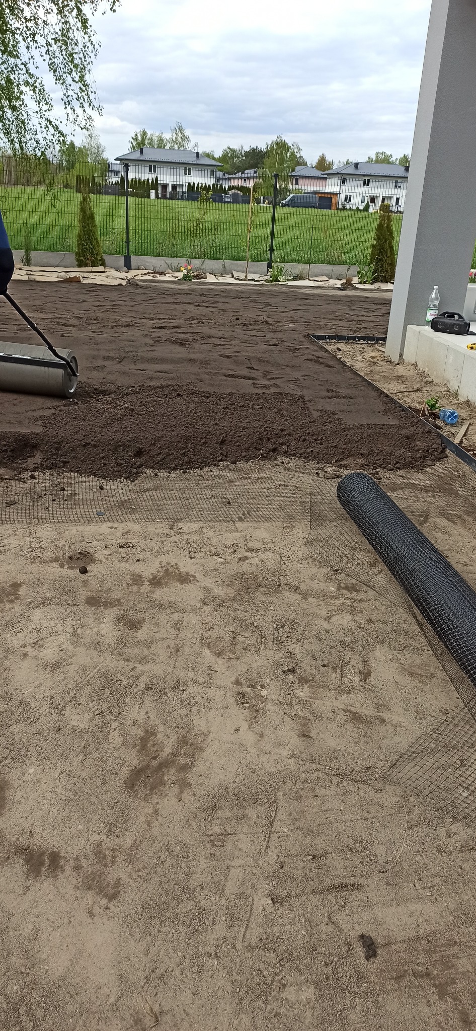 Przygotowanie terenu pod trawnik: rozkładanie siatki, wyrównywanie ziemi walcem. W tle domy jednorodzinne i ogrodzenie.