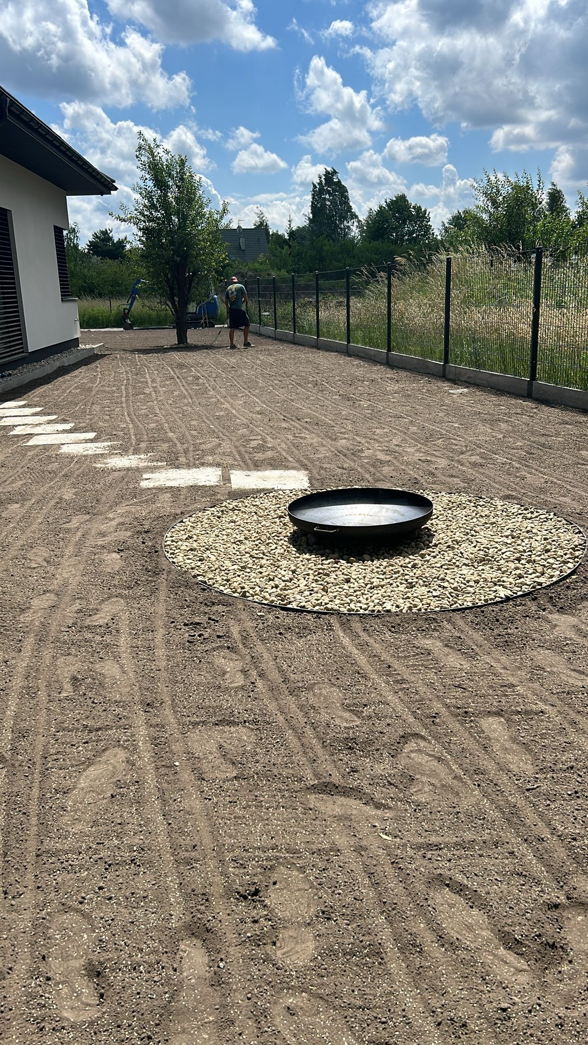 Przygotowany teren pod trawnik z okrągłym paleniskiem z kamieni. W tle pracownicy wyrównują teren. Widoczne ślady po maszynach.