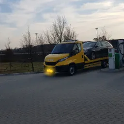 Żółta laweta Iveco z czarnym kombi na platformie, stojąca na stacji paliw z widocznymi dystrybutorami, w tle drzewa bez liści i pochmurne niebo.