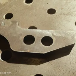 Surowy, metalowy element z dwoma okrągłymi otworami leżący na perforowanym stole spawalniczym w warsztacie.