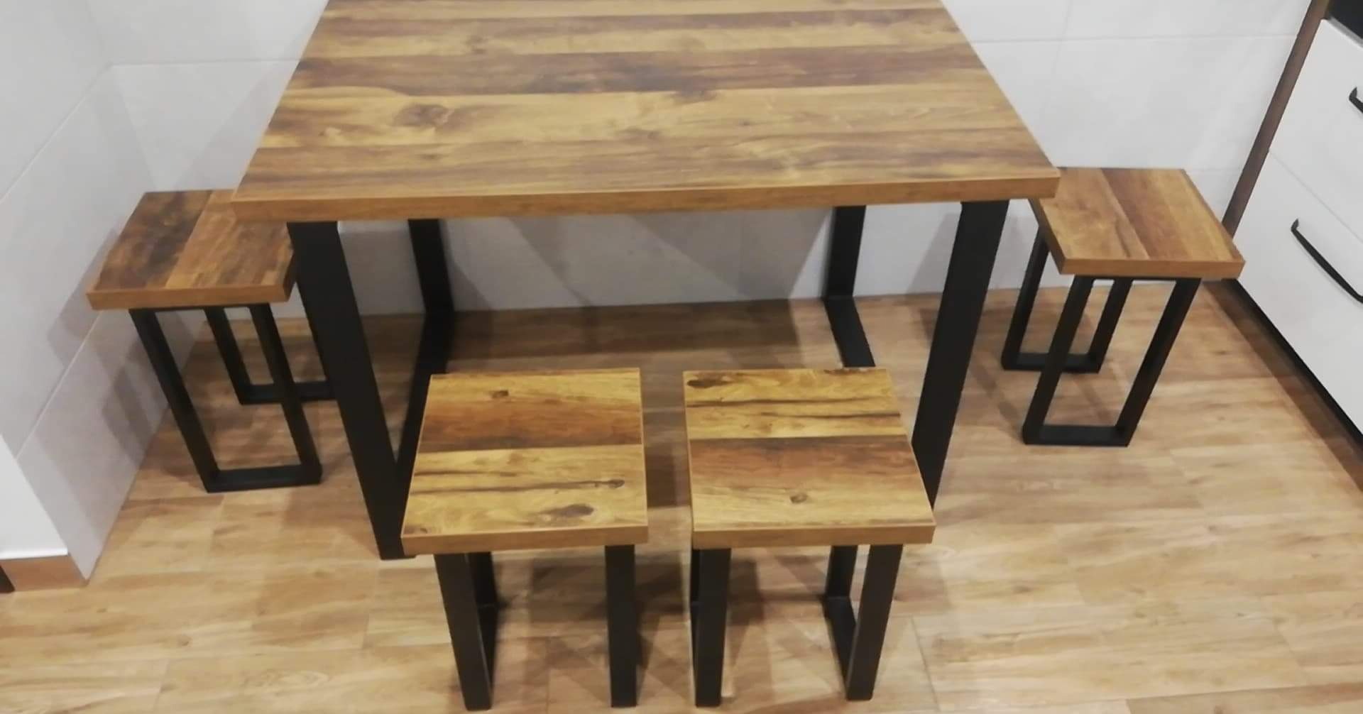 Kwadratowy stół z blatem z drewna o ciepłym odcieniu i czterema dopasowanymi stołkami z czarnymi, metalowymi nogami w stylu industrialnym, ustawiony na podłodze imitującej drewno.