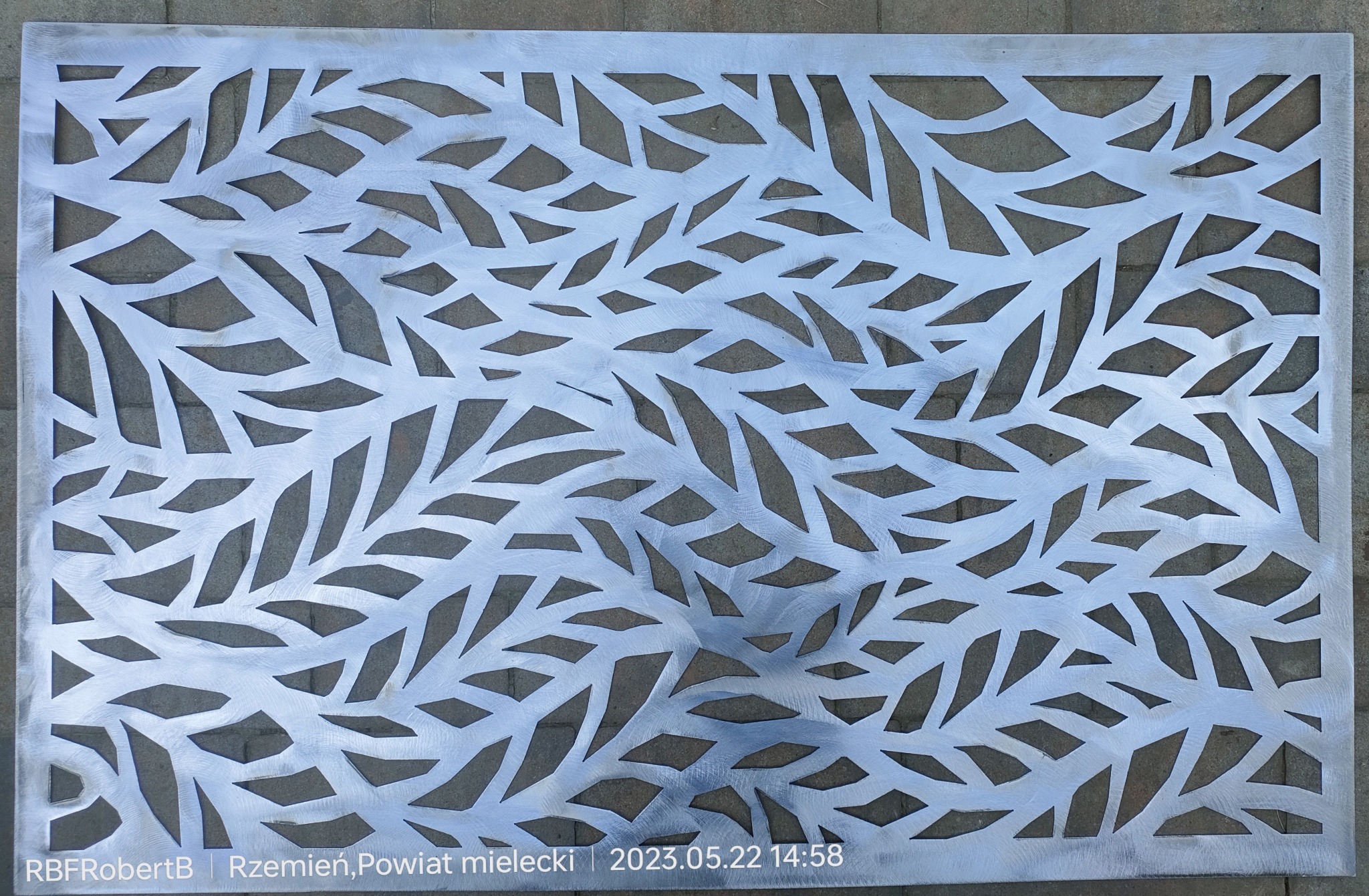 Metalowy panel dekoracyjny z wyciętym wzorem liści, widoczne ślady obróbki mechanicznej na powierzchni.