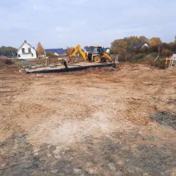 Żółta koparko-ładowarka przygotowuje teren pod fundament domu jednorodzinnego z widoczną izolacją przeciwwilgociową, w tle zabudowa mieszkalna.