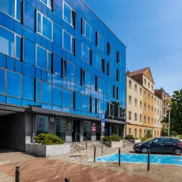 Nowoczesny biurowiec z niebieską szklaną fasadą odbijającą błękitne niebo, z parkingiem dla niepełnosprawnych i tabliczką z numerem 53 nad wejściem.