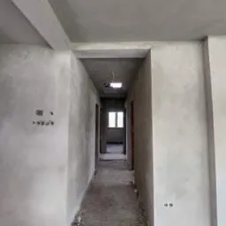 Korytarz w surowym stanie, ściany pokryte szarym tynkiem, widoczne puszki elektryczne, podłoga betonowa, perspektywa w głąb z oknem na końcu.