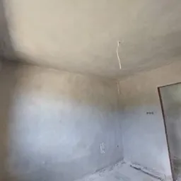 Surowe, szare ściany w pokoju z widocznym przewodem elektrycznym zwisającym z sufitu i niedokończonym otworem drzwiowym, ukazujące etap przygotowania do dalszych prac wykończeniowych.