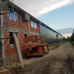 Budynek w trakcie ocieplania, z widocznym rusztowaniem, podnośnikiem i nowymi oknami, częściowo otynkowany, z zamontowaną klimatyzacją na elewacji.