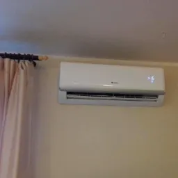 Biały klimatyzator marki Gree zamontowany na beżowej ścianie w pokoju, widoczne zasłony i karnisz.