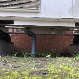 Klimasko Robert Szmit - Szara rura odprowadzająca skropliny z białej jednostki zewnętrznej, zamontowanej na metalowej konstrukcji wspartej na betonowych podstawach, widok z poziomu gruntu, zarośniętego mchem i trawą.