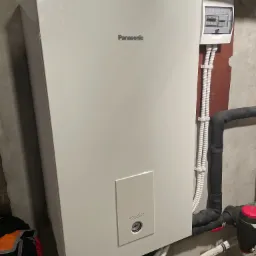 Biała jednostka wewnętrzna z logo Panasonic Aquarea, zamontowana na szarej ścianie w kotłowni, widoczne rury instalacyjne i skrzynka elektryczna.