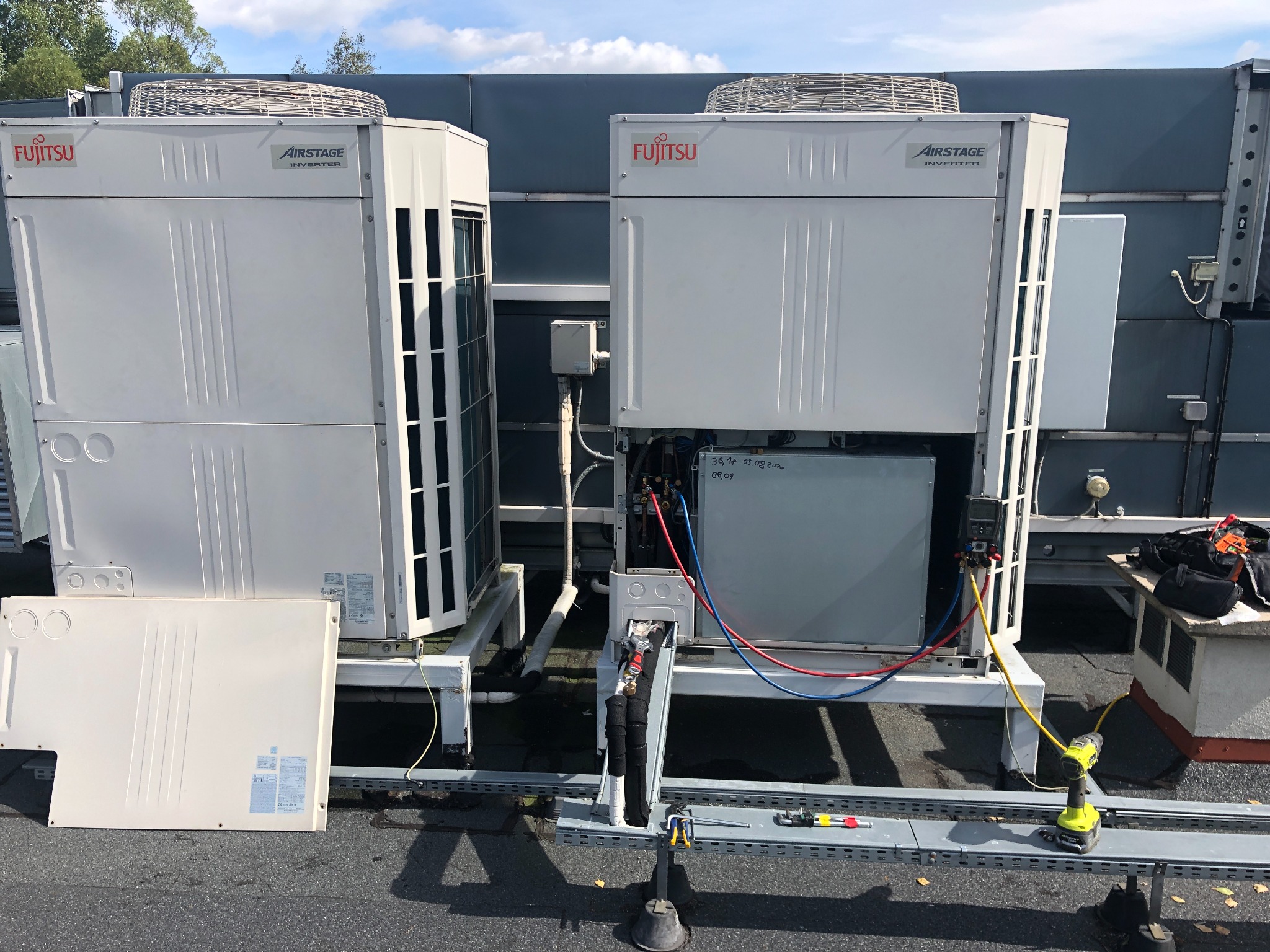 Dwa urządzenia klimatyzacyjne Fujitsu Airstage Inverter na dachu budynku, jeden z otwartym panelem dostępowym, widoczne przewody i narzędzia serwisowe, zarys nieba w tle.